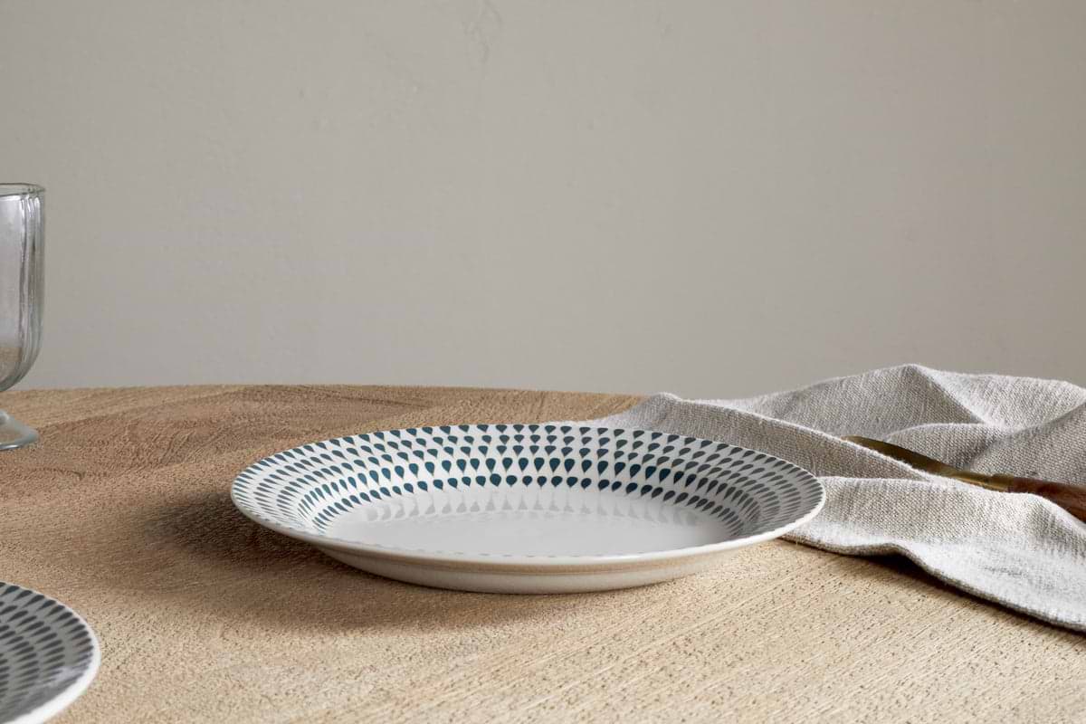 A modern dining scene: the nkuku Juniper Drop Side Plate in Cream & Juniper, featuring a blue dotted pattern, sits on a wooden table next to a beige napkin and a clear glass; the background is plain and neutral.