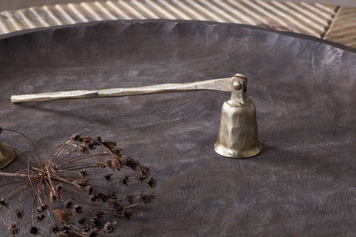 The nkuku Jyoti Candle Snuffer in antique brass, featuring a textured handle, sits on a dark metal tray beside dried botanicals—an elegant gift for candle lovers against a neutral, ridged background.