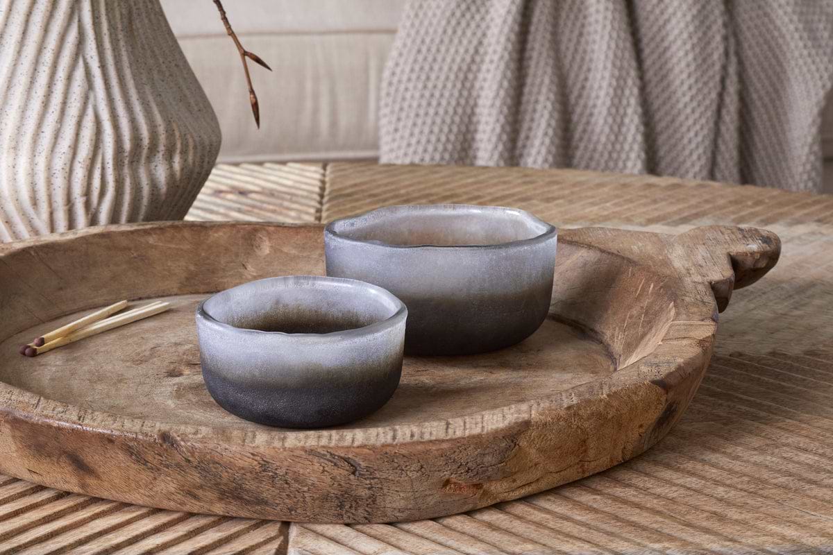 The nkuku Janka Tealight Holders - Smoked Charcoal (Set of 2) sit on a rustic wooden tray with matches, while a ceramic vase and knitted fabric in the background add to the cozy decor.