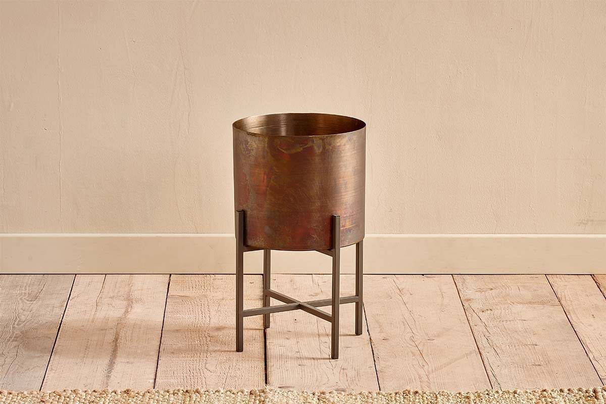 The Juoni Iron Planter by nkuku features a cylindrical, bronze-hued design on a black metal four-leg stand with crossbars, set on light wood flooring against a beige wall. A woven rug borders the scene—ideal for showcasing house plants.