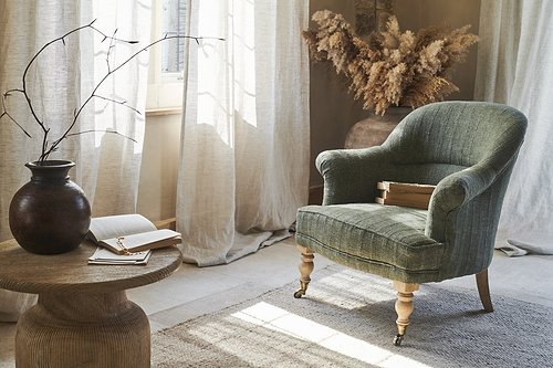 A Kiana Cotton Armchair in Moss by nkuku sits on a rug beside a round hand-carved mango wood table topped with a dark vase, branch, open book, and notebook. Light curtains and dried grasses complete the softly lit, neutral room.