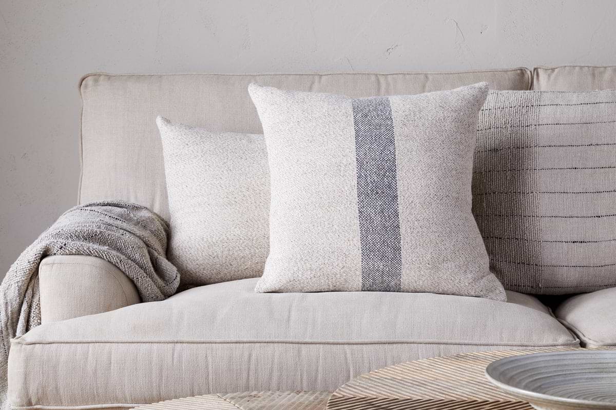 A beige sofa features the nkuku Kallu Wool & Cotton Cushion Cover in natural, a textured cream cushion, and a gray throw blanket. A light background wall and woven tray on the table finish this serene setting.