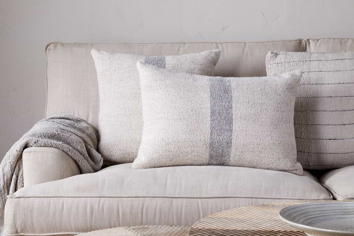 A beige sofa with nkuku’s Kallu Wool & Cotton Cushion Cover - Natural and three neutral throw pillows sits against a light wall, a gray knit blanket over the armrest. A round, light wood table appears in the foreground.