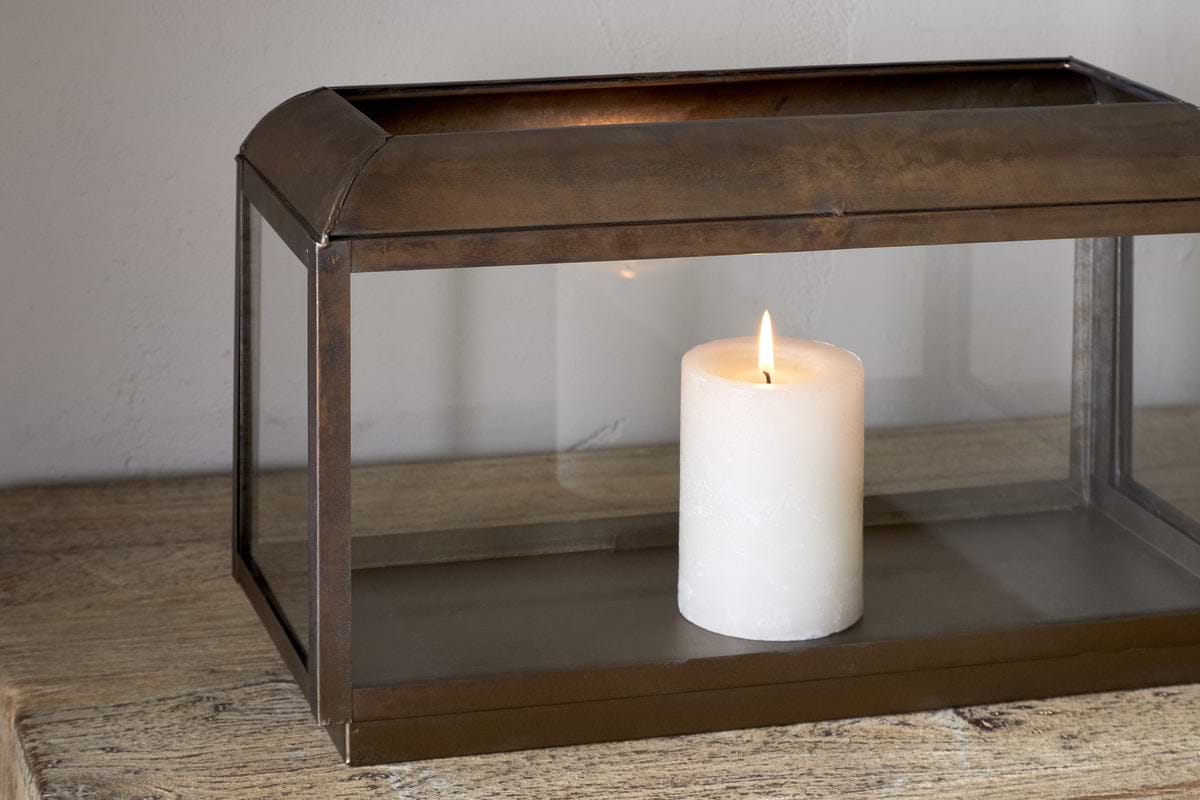The Kiwanda Tall Lantern - Antique Brass by nkuku features a white pillar candle burning inside its rectangular metal and glass frame, adding timeless charm to any wooden surface and creating elegant candlelight against light-colored walls.