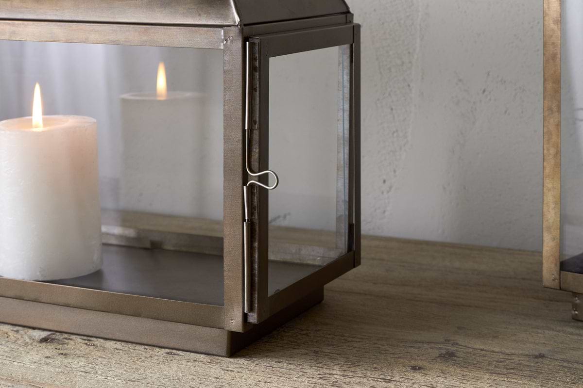 The Kiwanda Wide Lantern by nkuku, crafted in antique brass with glass panels, sits on a wooden surface against a light textured wall, bringing charm to candlelight décor.