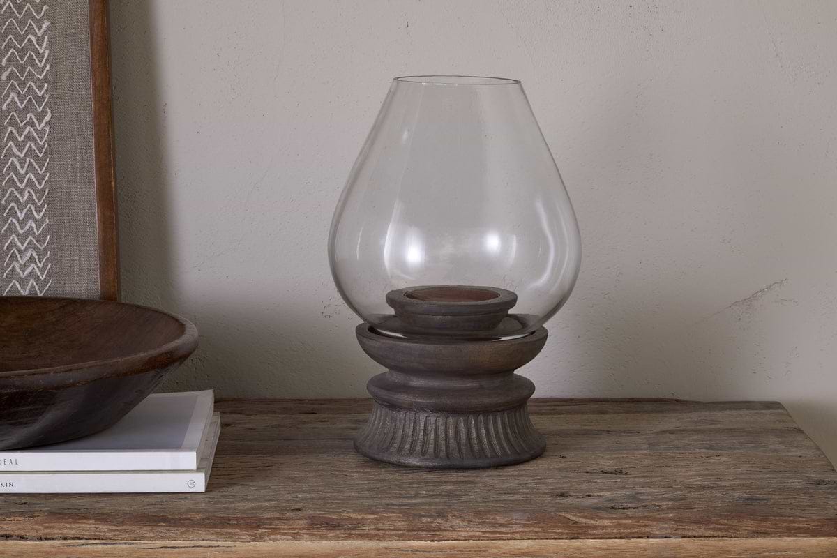 The nkuku Kawani Mango Wood Lantern in Dark Ash rests on a rustic table beside a shallow wooden bowl and white books, perfect for displaying pillar candles in style.
