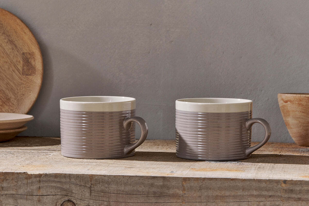 The nkuku Kai Mugs - Grey (Set of 2) feature ribbed textures, taupe bodies, and cream rims. They are displayed side by side on rustic wood, with blurred wooden plates and a bowl in the background against a light gray wall.