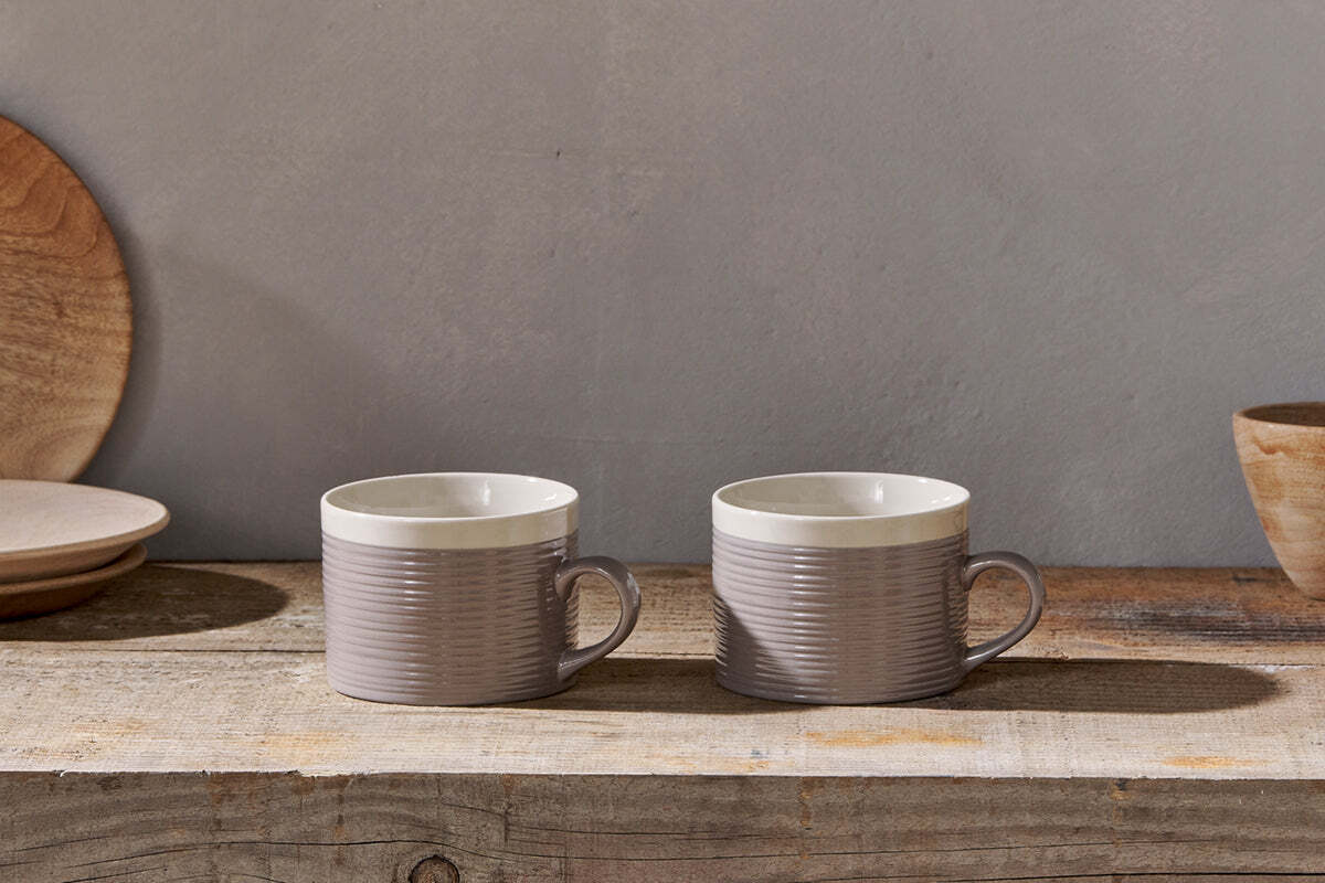 The Kai Mugs - Grey (Set of 2) by nkuku, featuring contemporary ribbed detail, rest on a rustic wooden surface with light brown wooden plates and a bowl behind them, all softly lit for a cozy ambiance.