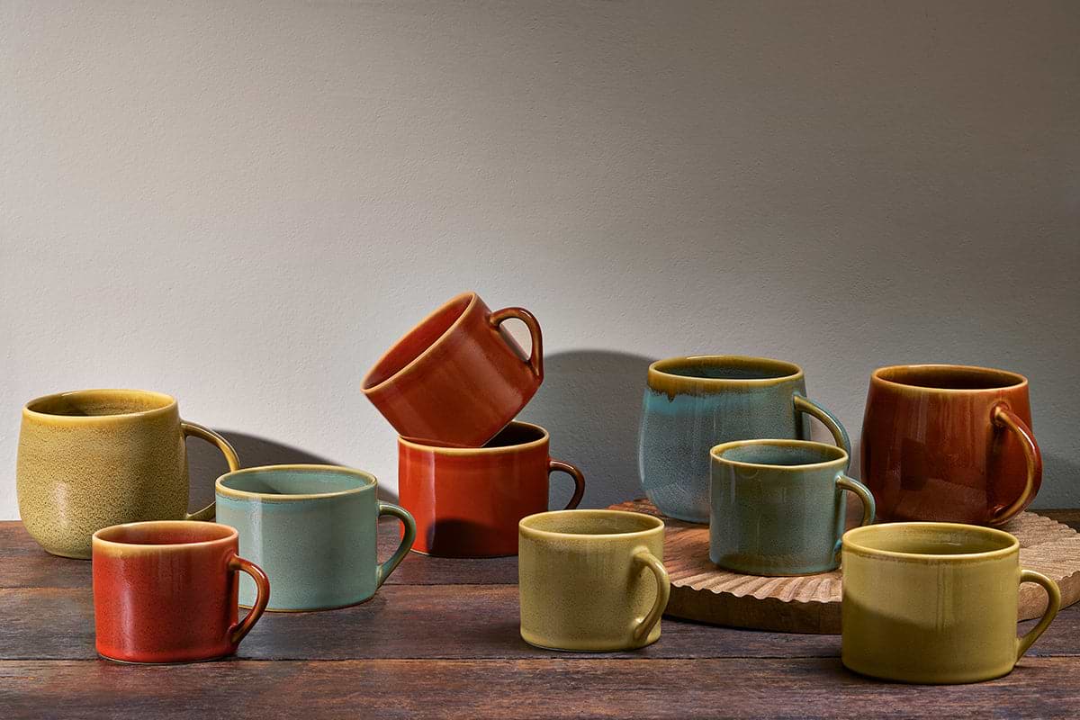 Ten ceramic mugs, featuring the nkuku Kalini Mug - Green (Extra Large), in earthy shades of yellow, orange, blue, and green, are displayed on a wooden tray and surface. Some mugs are stacked or leaning, casting soft wall shadows.