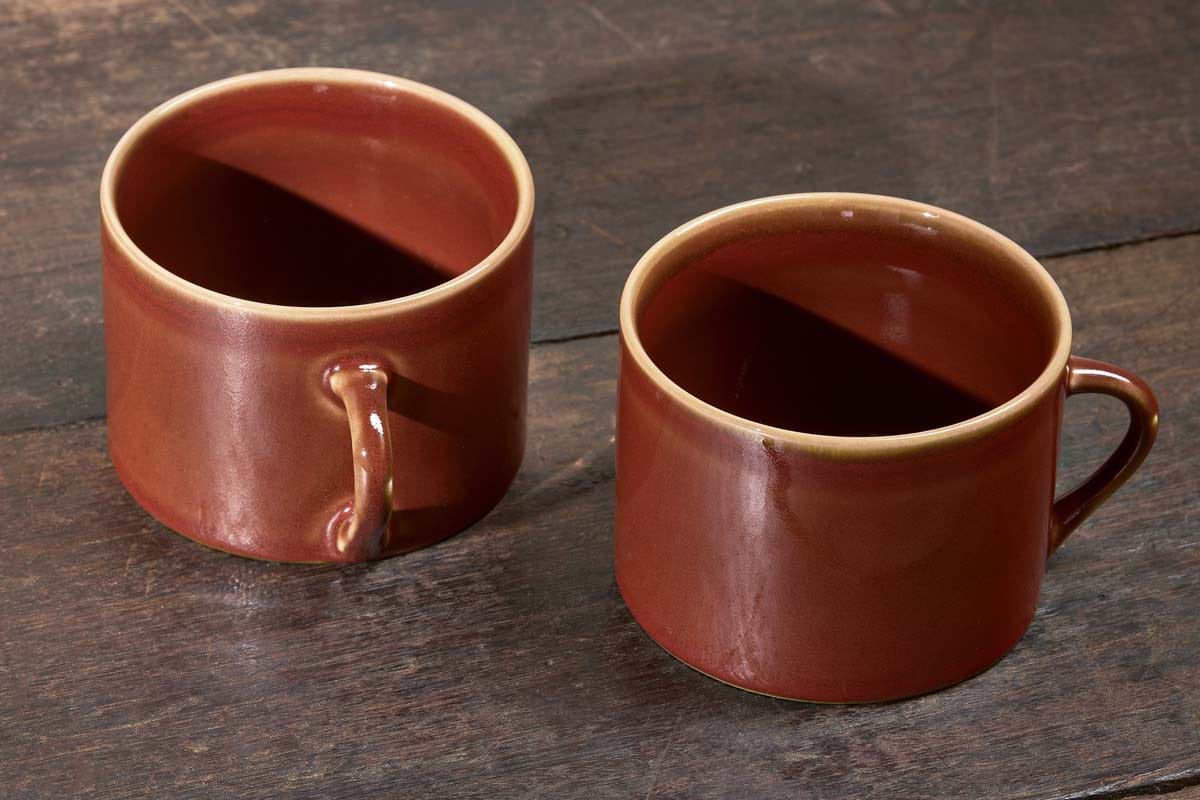 A set of two large, kiln-fired Kalini Mugs - Amber by nkuku with deep red bodies and light brown rims rests on a dark, weathered wooden surface; the left mug sits closer and faces left, the right mug faces right.