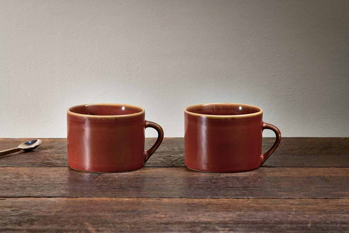 Two kiln-fired nkuku Kalini Mugs - Amber - Large (Set of 2) with handles rest side by side on a rustic wooden table. A small metal spoon with a dark handle lies to the left, set against a light beige background for a simple, cozy vibe.