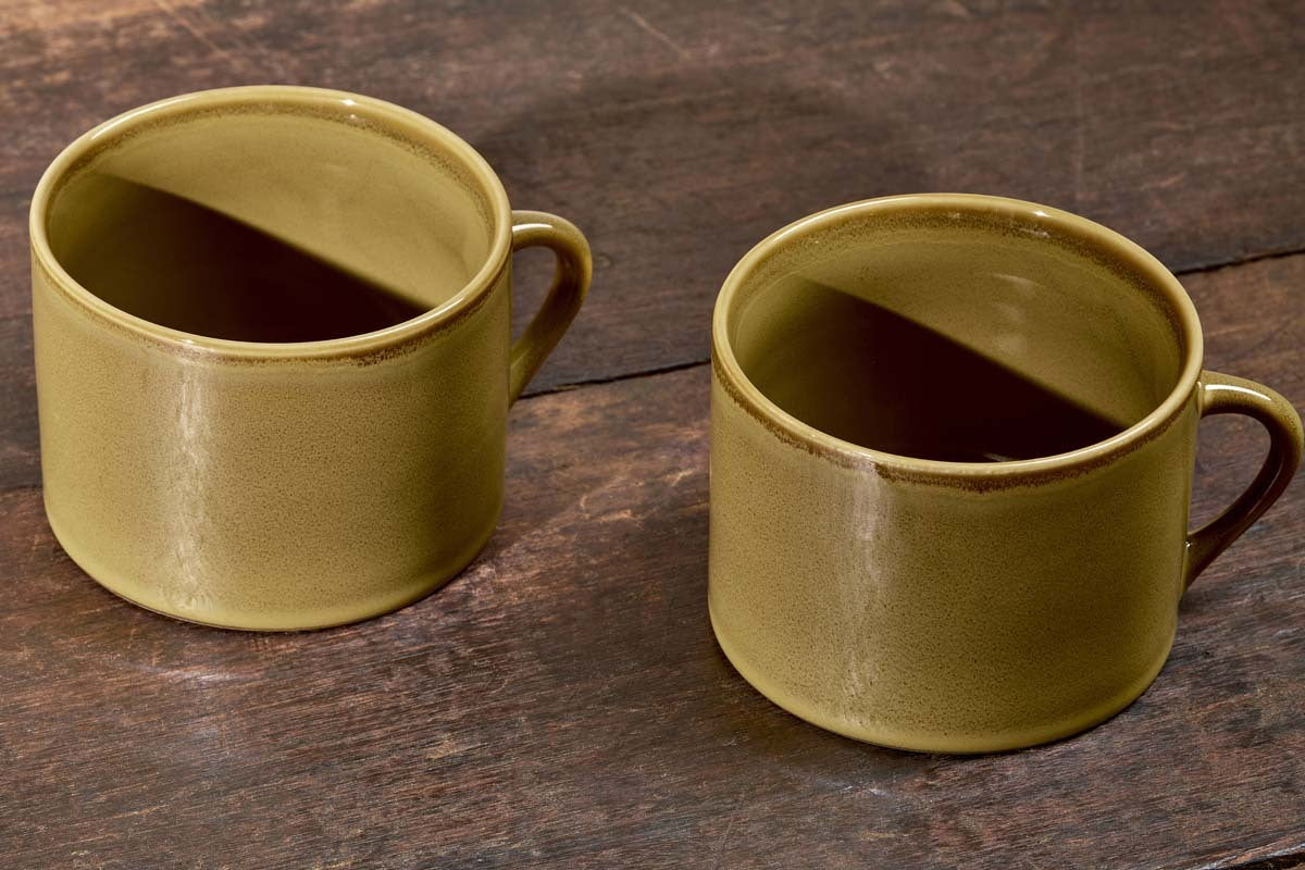 The Kalini Mugs - Green - Large (Set of 2) by nkuku, featuring a textured olive-green glaze and sturdy handles, rest on a dark wooden surface with shadows falling inside each mug.