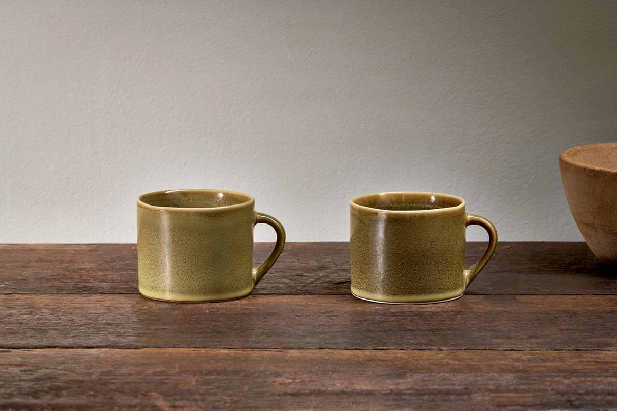 A set of two small green Kalini mugs by nkuku with handles are placed side by side on a rustic wood surface, with a brown ceramic bowl partly visible on the right and a plain, light-colored wall in the background.