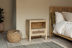 A cozy bedroom features the nkuku Kanz Mango Wood Bedside Table with an open shelf and handwoven cane drawer, beside a wooden bed with white bedding. A round, woven basket rests on the light wood floor, enhancing the calm, neutral decor.