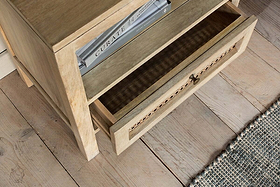 The nkuku Kanz Mango Wood Bedside Table with an open drawer stands on a matching plank floor, holding the book “Curate” on its shelf. A blue and white textured rug appears partially in the lower right corner.