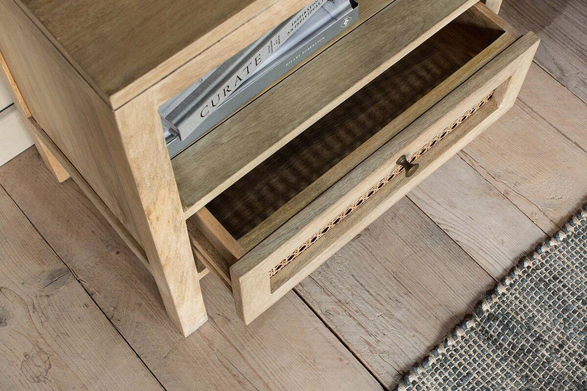 The nkuku Kanz Mango Wood Bedside Table with an open drawer stands on a matching plank floor, holding the book “Curate” on its shelf. A blue and white textured rug appears partially in the lower right corner.