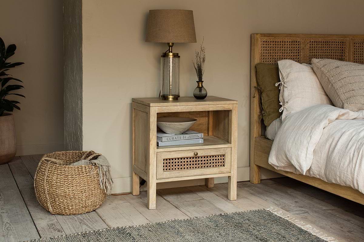 The nkuku Kanz Mango Wood Bedside Table, featuring a handwoven cane drawer and open shelf, offers stylish and practical bedside storage next to a rattan headboard and linen bedding.