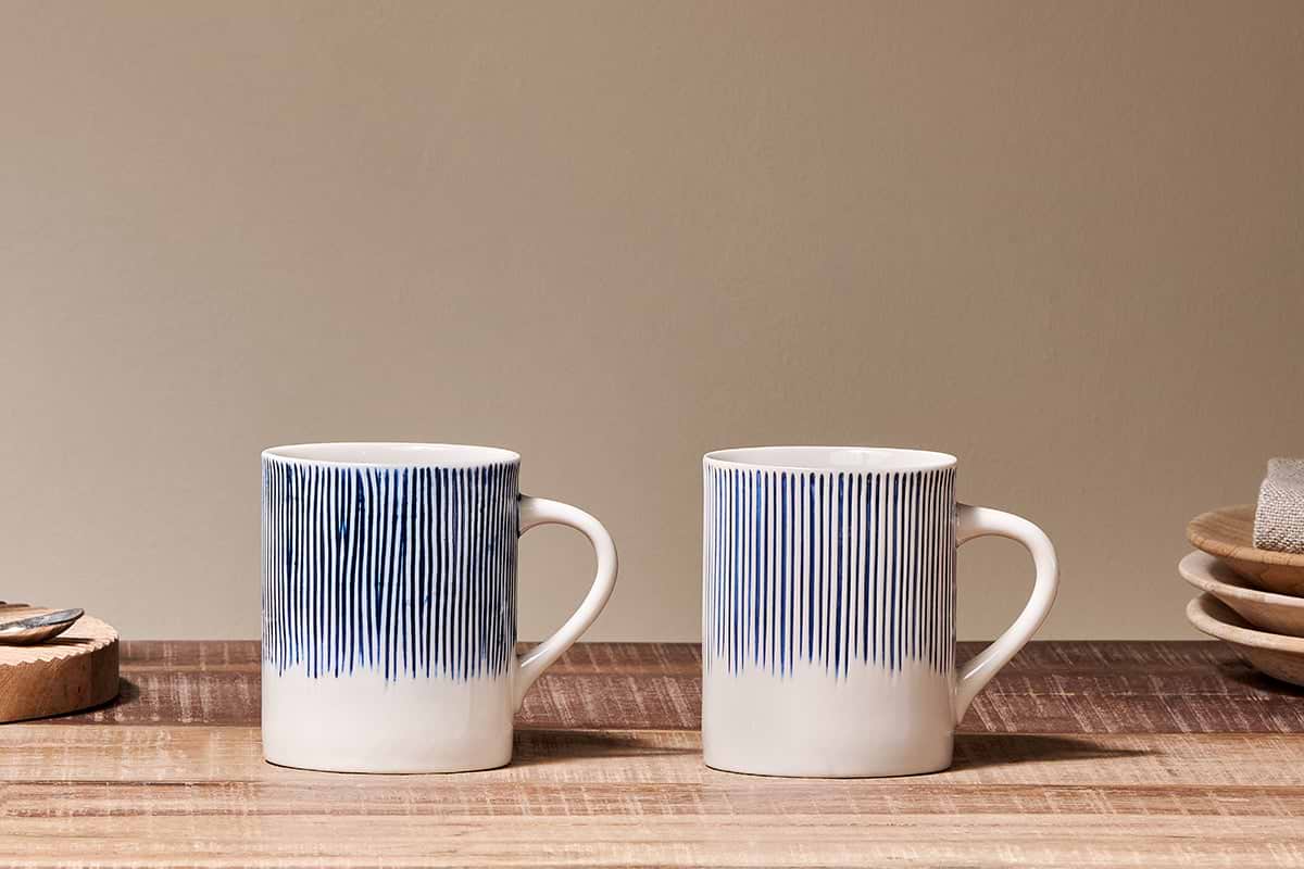 Two white nkuku Karuma Ceramic Mug - Large (Set of 2), featuring vertical blue lines, sit side by side on a wooden surface, with handmade mugs, stacked plates, a napkin, and a cork coaster in the neutral beige background.