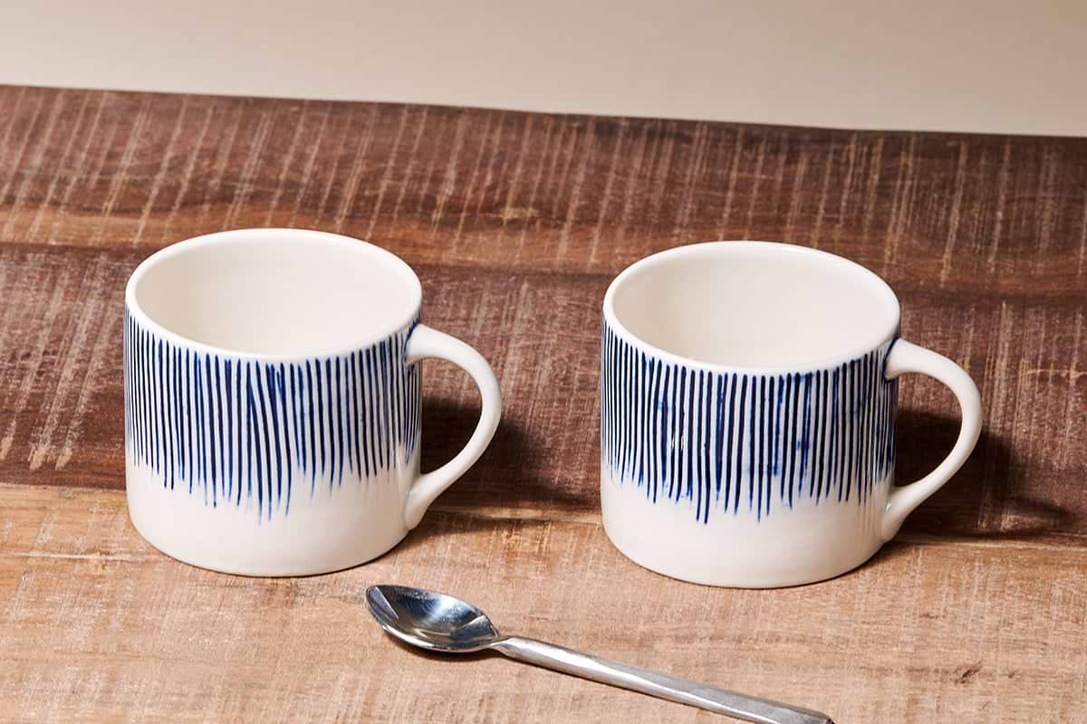 The Karuma Ceramic Mug - Small (Set of 2) by nkuku features handmade Vietnamese-style ceramics with vertical blue lines on the upper half, shown on a rustic wooden surface with a metal spoon and a plain beige background.