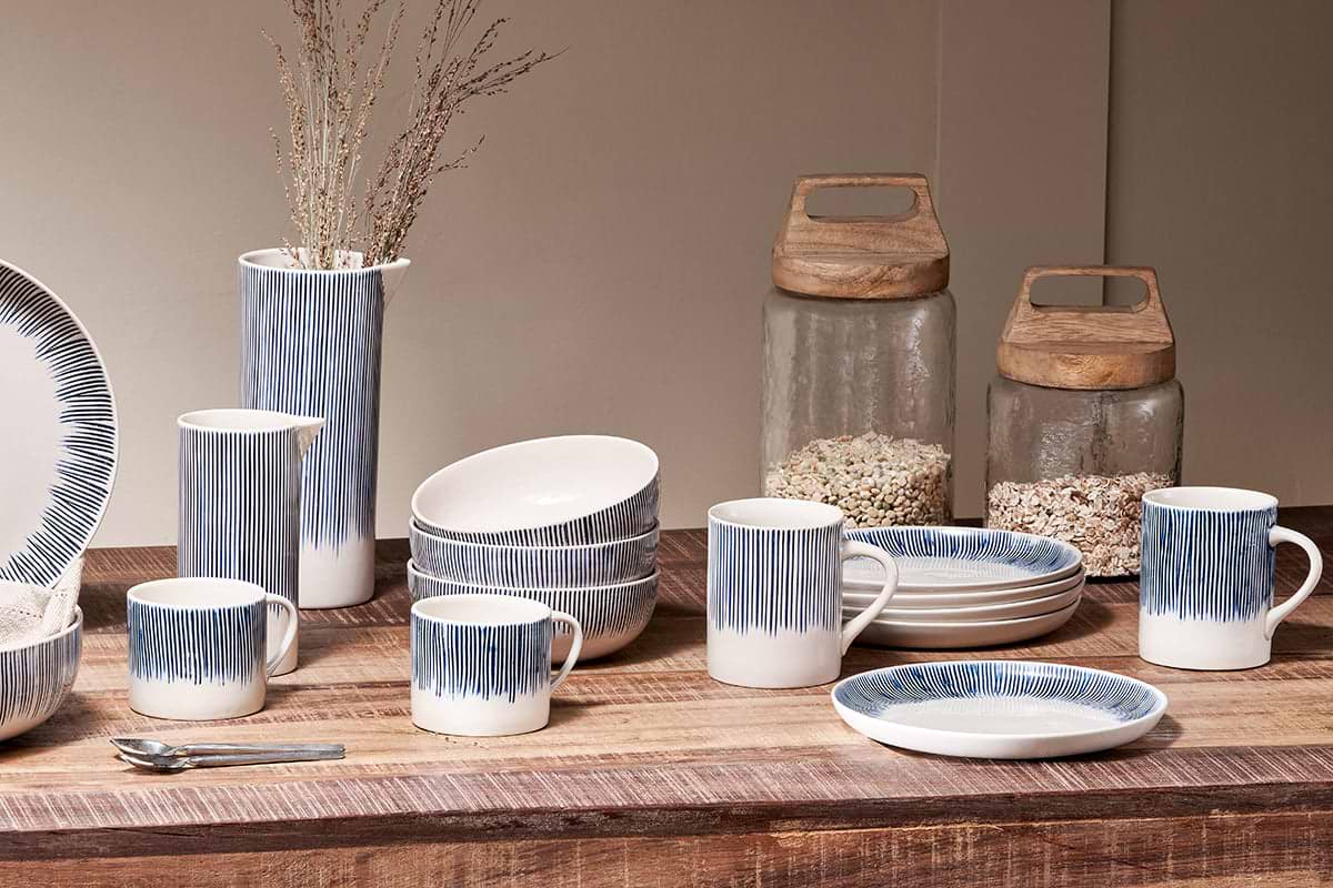The Karuma Ceramic Mug - Small (Set of 2) by nkuku features white Vietnamese ceramics with blue vertical stripes, handmade design, and pairs well with plates, bowls, and a matching pitcher. Two glass jars with wooden lids sit in the background.
