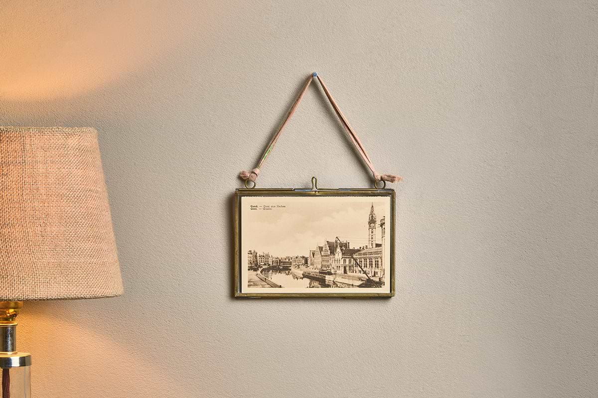 A Kiko Brass Frame by nkuku, displaying a vintage European cityscape postcard, hangs on a light beige wall by a recycled sari tie. Next to it, a textured lampshade softly illuminates the space with a warm glow.