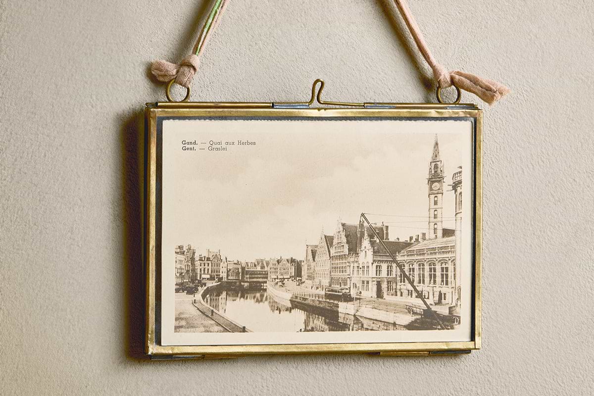 A vintage black-and-white canal scene is displayed in the Kiko Brass Frame by nkuku, which hangs on a beige wall with a pink recycled sari tie. The upper left of the photo reads: “Gent. Quai aux Herbes. Gand.—Graslei.”.
