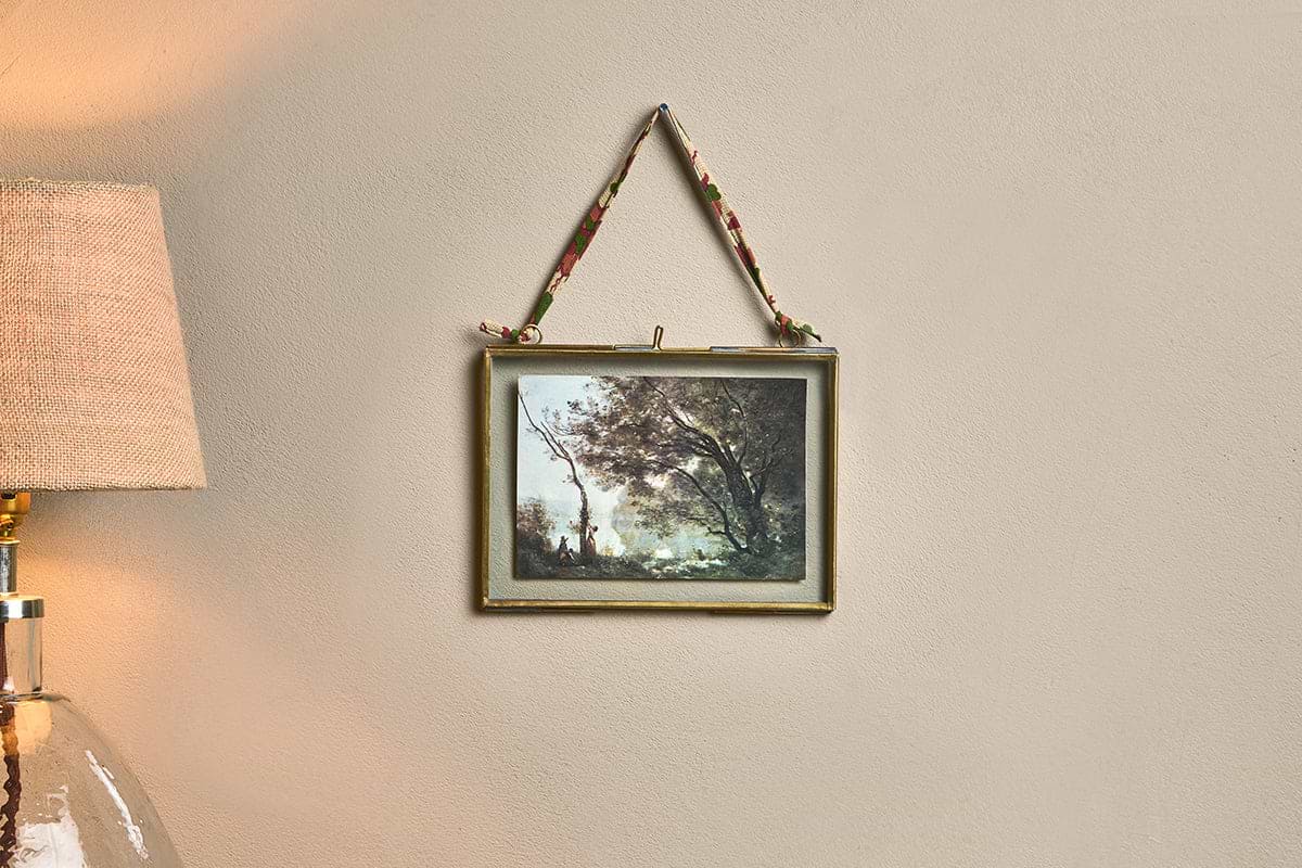 A Kiko Brass Frame by nkuku, showcasing a landscape painting with trees and a river, hangs from a recycled sari tie. To the left are part of a beige lampshade and glass lamp, while the plain light wall creates a cozy atmosphere.