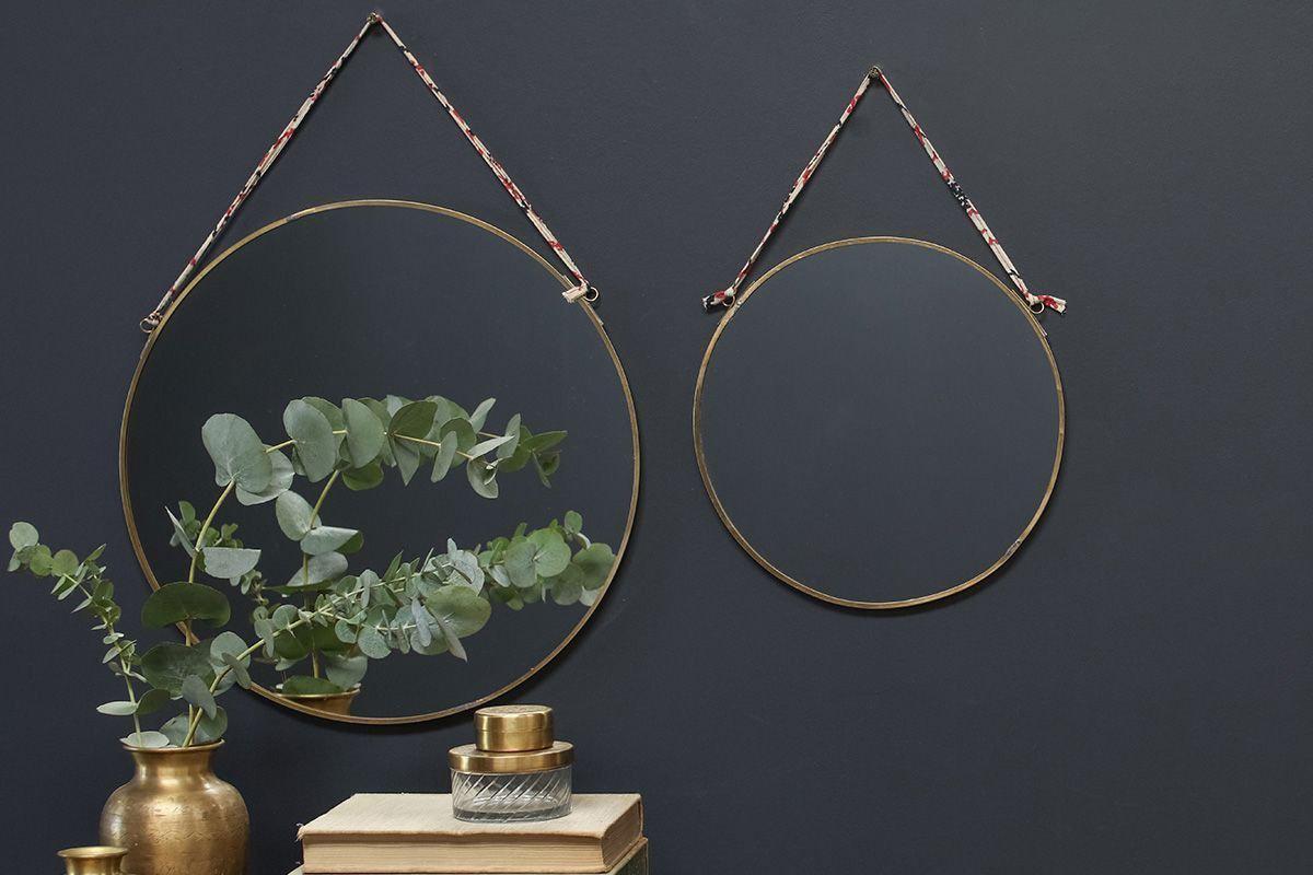 Two nkuku Kiko Round Mirrors - Brass with recycled sari tie straps hang on a dark wall. Below, a gold vase with green eucalyptus, a small gold jar, and a ribbed glass container sit atop stacked beige books.