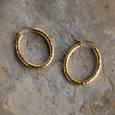 The Labani Earrings - Gold by nkuku, featuring a textured hoop design, are displayed on a gray stone surface, highlighting their timeless elegance.