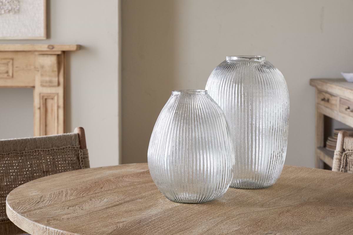 Two Linsy Ribbed Glass Vases by nkuku in clear, each a different height, sit on a round wooden table. Natural, neutral tones surround them, with a woven chair, light wood furniture, and soft beige walls in the background.
