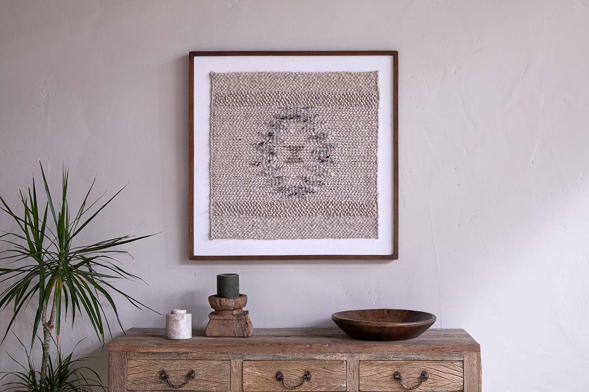 A rustic wooden dresser with three drawers holds a potted plant, candles, and a wooden bowl. Above, the nkuku Lena Textured Wall Art in Off White adds artisanal charm to the light beige wall.