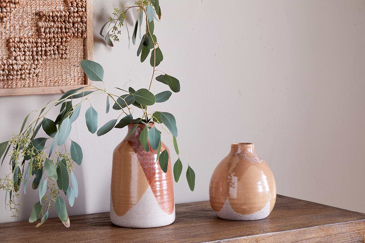 The nkuku Lucia Ceramic Vase - Terracotta Mix features glossy, earth-toned geometric patterns and holds eucalyptus branches. It sits alongside another vase on a wooden surface, near a woven wall hanging against a light wall.