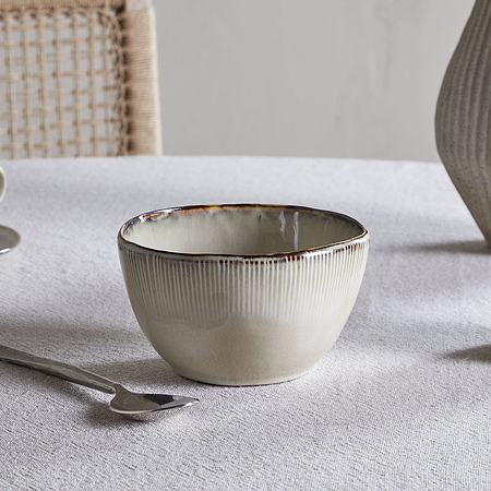 The nkuku Malia Breakfast Bowl - Cream, featuring a reactive glaze and organic shape, rests on a textured white tablecloth alongside a metal spoon, cup and saucer, small pitcher, ribbed vase, and a woven chair in the background.