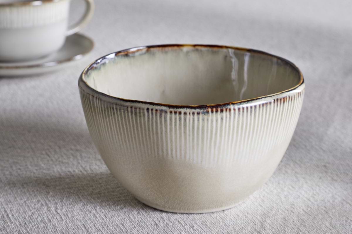 A cream Malia Breakfast Bowl by nkuku, featuring a gently irregular shape and vertical ridges, sits on a light textured surface. In the background, a matching cup and saucer with a subtle reactive glaze are softly blurred.