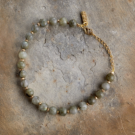 The Marai Bracelet - Labradorite by nkuku showcases round greenish-gray labradorite stones with gold spacers, a gold chain and clasp, elegantly presented on a textured, earth-toned background.