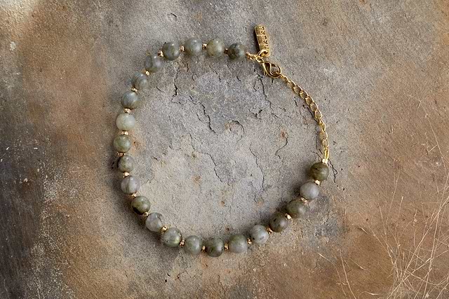 The Marai Bracelet - Labradorite by nkuku showcases round greenish-gray labradorite stones with gold spacers, a gold chain and clasp, elegantly presented on a textured, earth-toned background.
