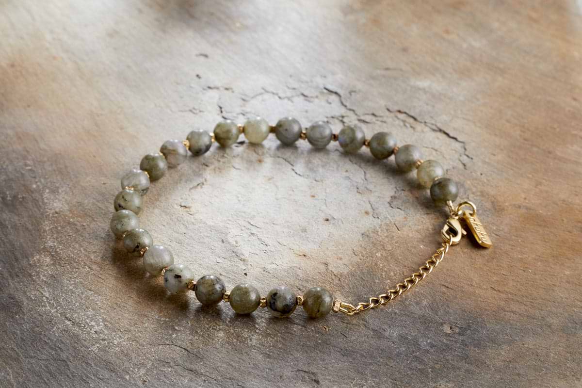 The Marai Bracelet - Labradorite by nkuku features round, mottled greenish-grey labradorite stones and a gold chain clasp, elegantly presented on a textured wooden surface.