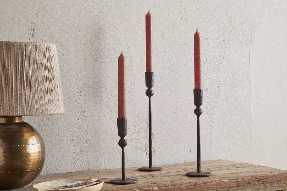 Three red taper candles in nkuku’s Mawindi Candle Holders - Rust are displayed at different heights on a rustic wood surface beside a beige and brass lamp, all set against a textured light wall for an earthy decor accent.