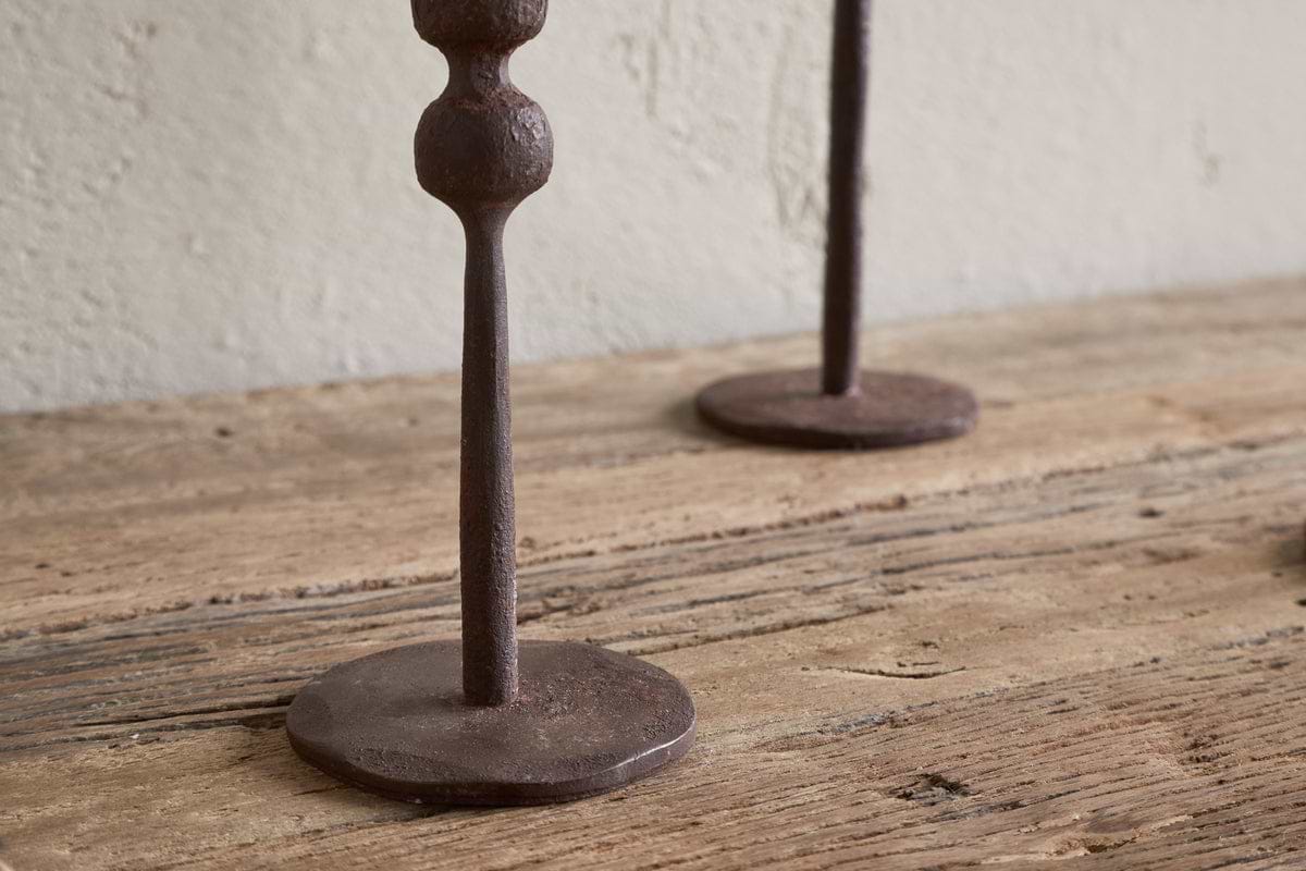 Two Mawindi Candle Holders in rust by nkuku sit on a wooden surface against a beige textured wall, their round bases and lower stems adding charm to earthy decor.