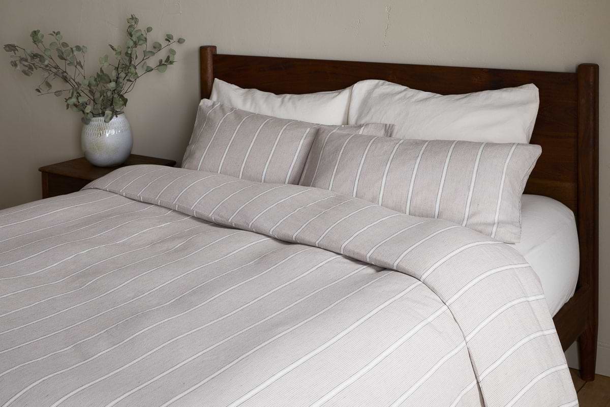 A neatly made bed featuring the nkuku Mora Hemp & Cotton Duvet Cover in Stone Grey, paired with two large pillows and a wooden headboard. A white vase with leafy branches sits on a bedside table against a neutral wall.