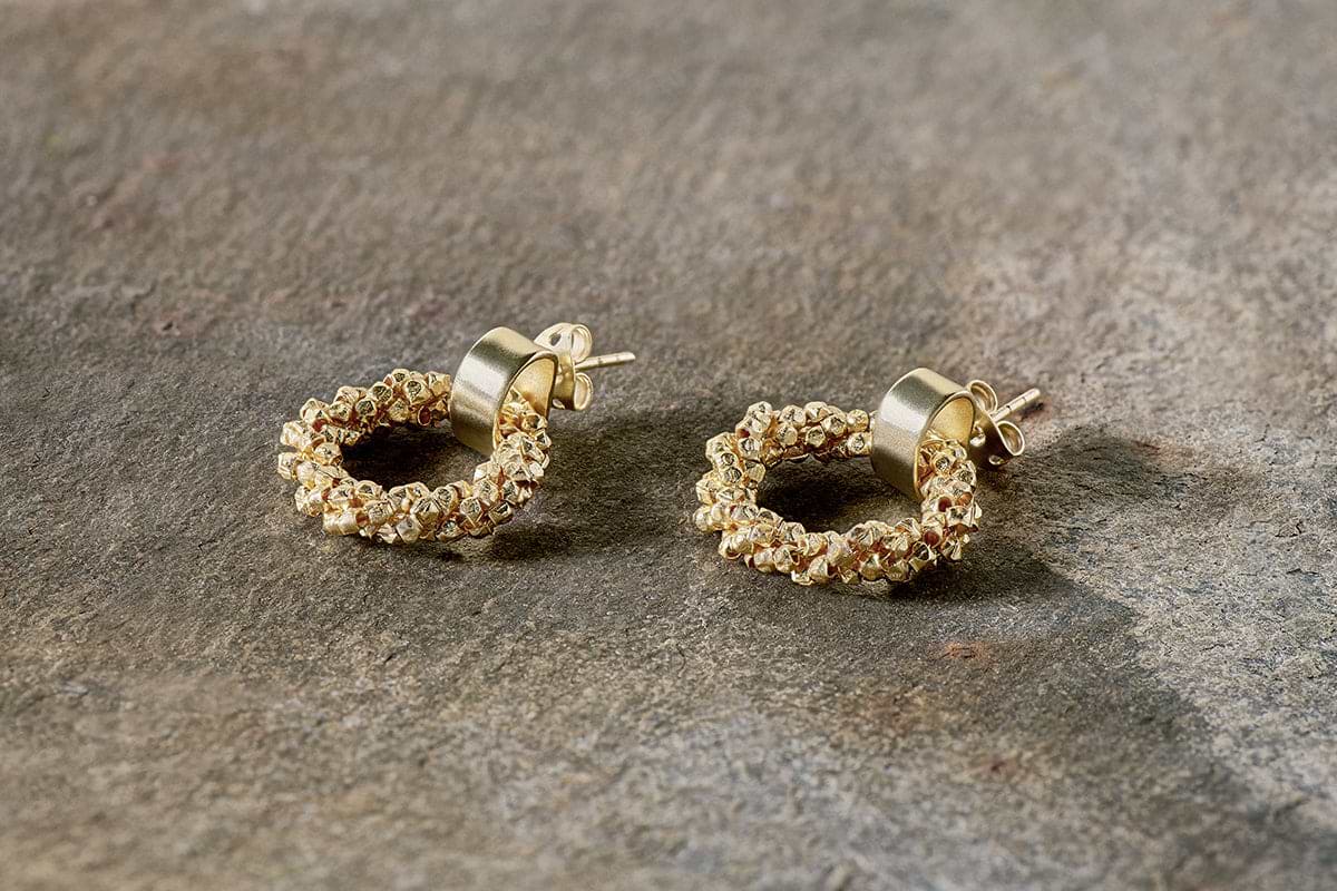The Mula Earrings - Gold by nkuku are small, gold-plated hoops with a textured beaded design. Hypoallergenic, they feature round posts and circular studs for secure fastening and are shown resting on a rough stone surface.