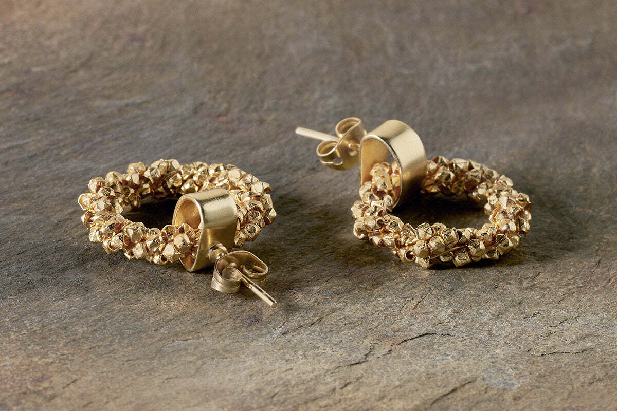The nkuku Mula Earrings - Gold feature textured circular hoops made of faceted brass beads clustered on smooth cylindrical bases. Hypoallergenic and ideal for sensitive ears, they are shown on a stone-like surface.