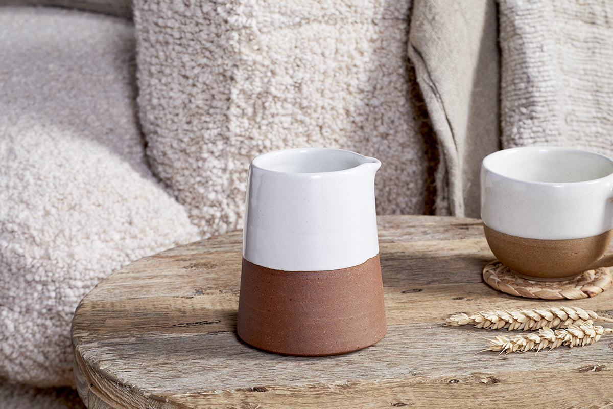 The nkuku Mali White and Terracotta ceramic pitcher sits on a rustic table beside the matching cup and wheat stalks. A textured beige sofa with a draped throw blanket creates a cozy backdrop.