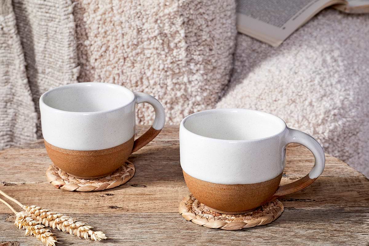 Two nkuku Mali White and Terracotta ceramic mugs rest on woven coasters atop a rustic wooden table. Paired with a soft throw and an open book, they create a cozy, inviting handmade pottery scene.