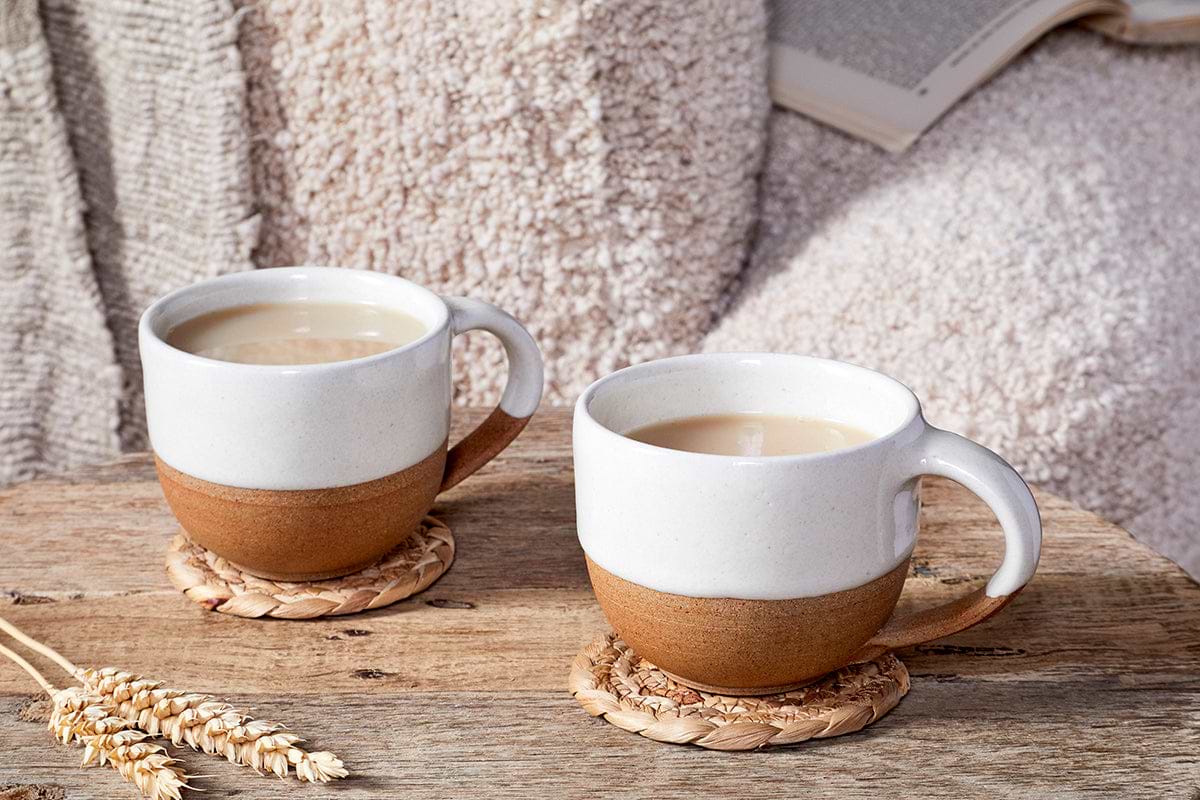 Two nkuku Mali White and Terracotta handmade mugs, filled with light brown liquid, rest on woven coasters atop a rustic wooden surface. A soft, textured blanket and an open book appear in the background.