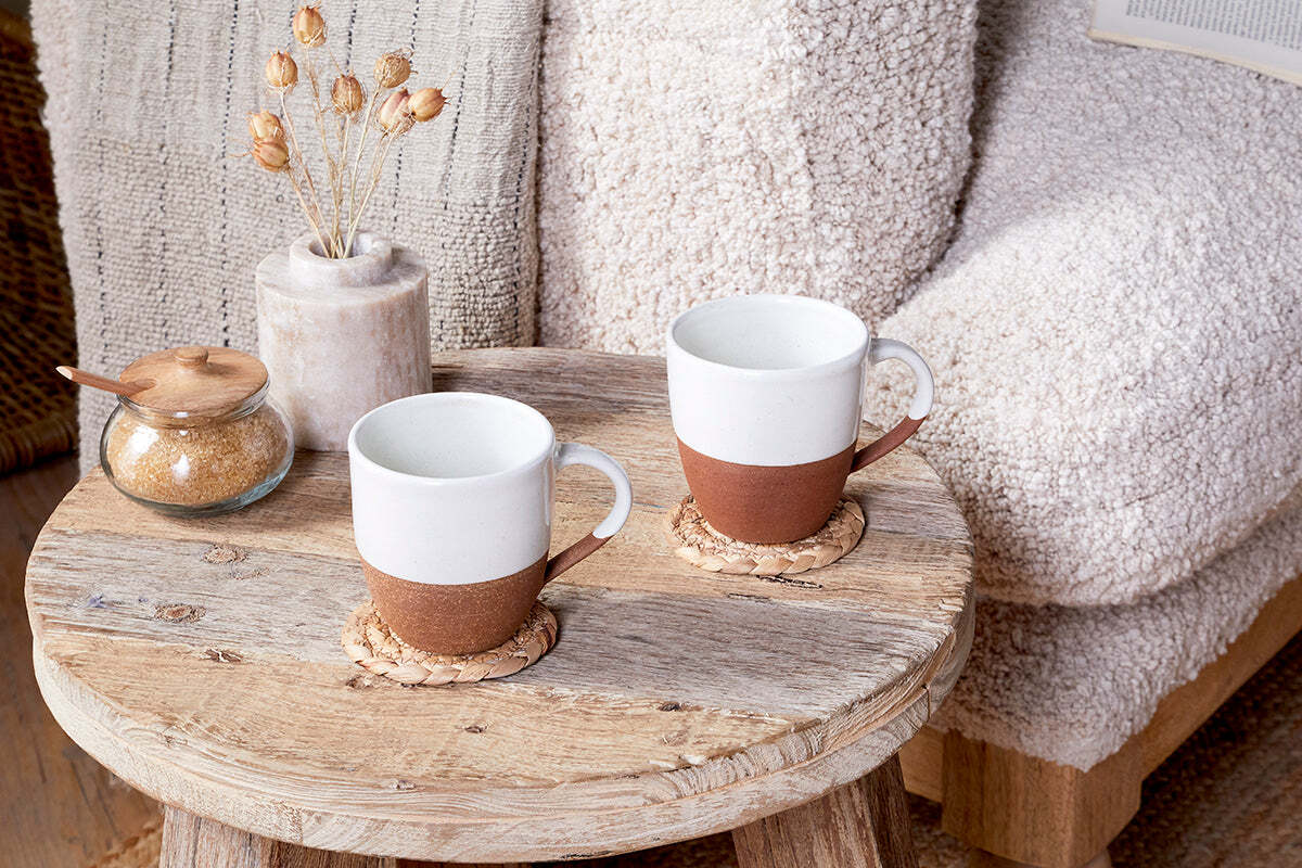 A rustic wooden coffee table holds two nkuku Mali White & Terracotta mugs on woven coasters, a jar of brown sugar with a wooden lid, and a small vase with dried flowers. In the background sits a textured beige armchair with a soft throw.