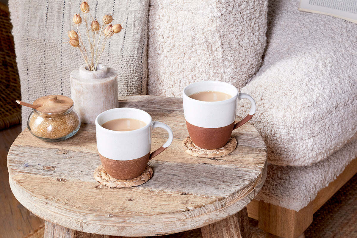 Two nkuku Mali White & Terracotta mugs with milky coffee rest on woven coasters on a rustic table, beside a brown sugar jar with a wooden lid and a ceramic vase of dried flowers. A textured beige armchair is partly visible.