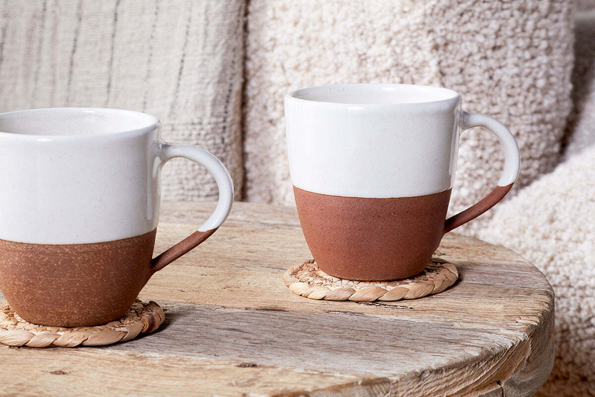 Two nkuku Mali White & Terracotta mugs with off-white glazed tops and handmade terracotta bases sit on a wooden table, each resting on a woven coaster.
