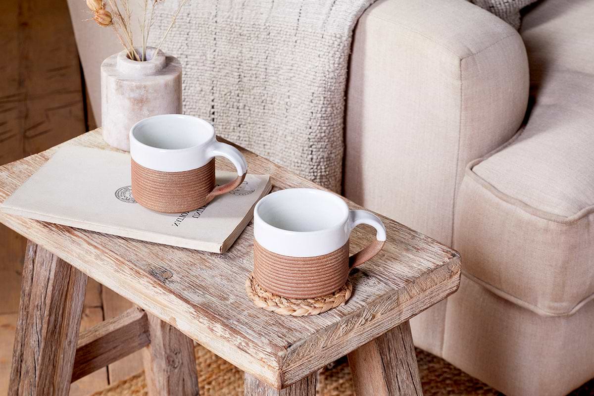 The nkuku Mali Ribbed Coffee Mug - White (Set of 2) adds artisan charm to your cozy corner when placed on a rustic stool beside a beige armchair with a textured throw blanket.