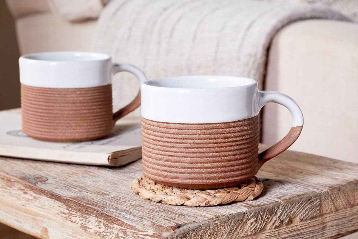 The nkuku Mali Ribbed Coffee Mug - White (Set of 2) features contemporary mugs with ribbed brown bases, displayed on a rustic table—one on a coaster, the other atop a book beside a beige blanket and sofa in the background.